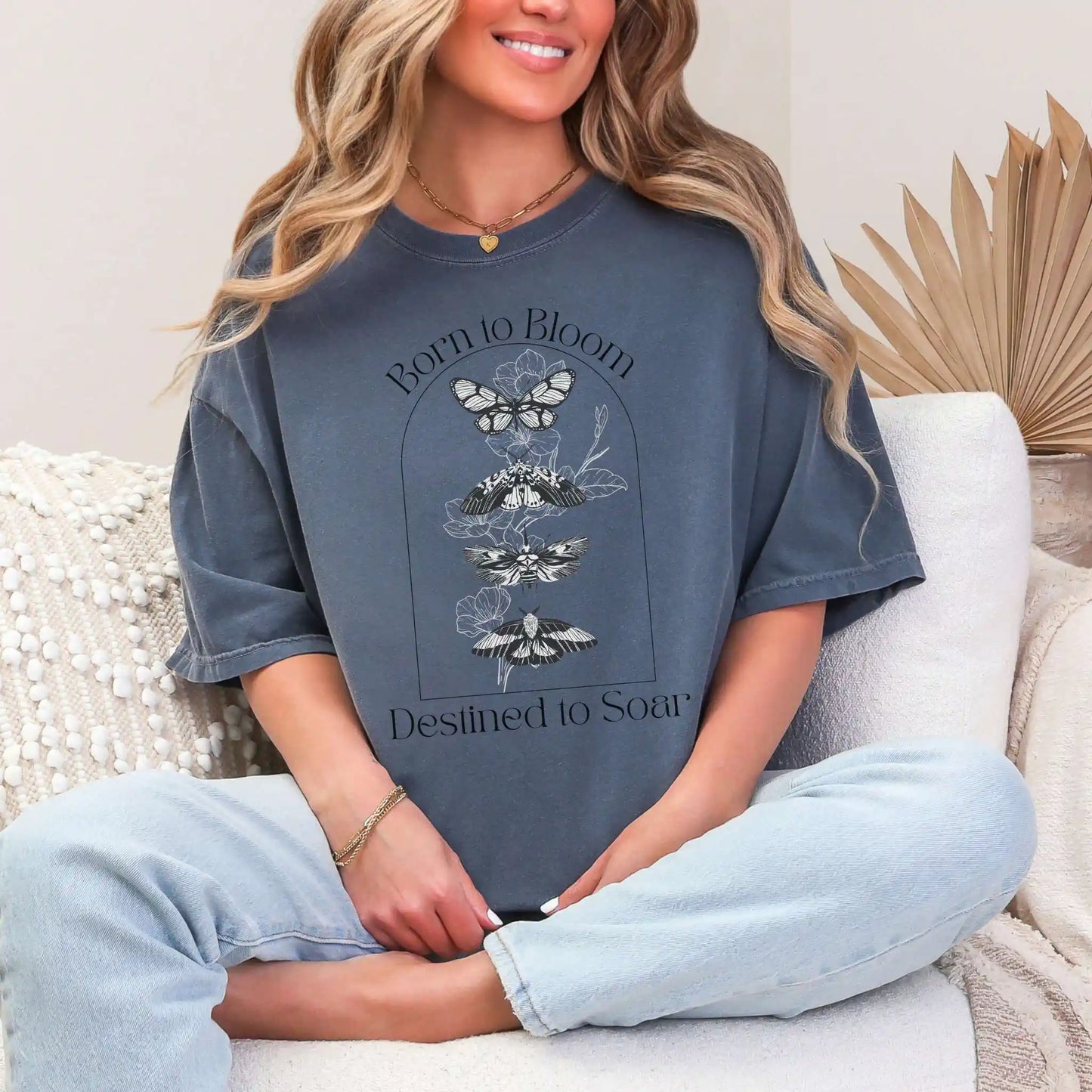 Woman wearing a blue oversized t-shirt with butterfly design and 'Born to Bloom, Destined to Soar' text, sitting on a white couch.