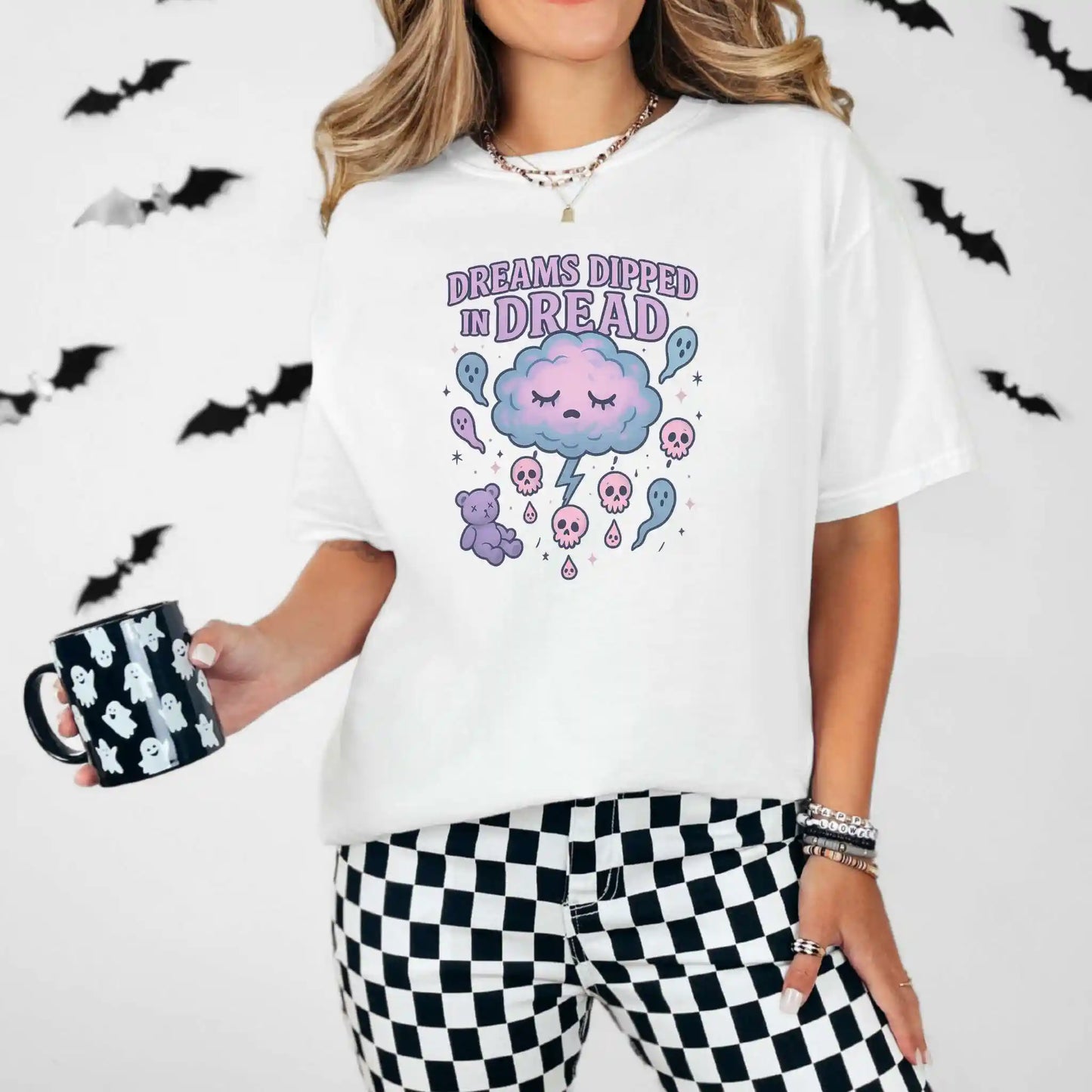Person wearing a white t-shirt with a "Dreams Dipped in Dread" graphic design with clouds and ghosts, holding a mug, surrounded by bats.