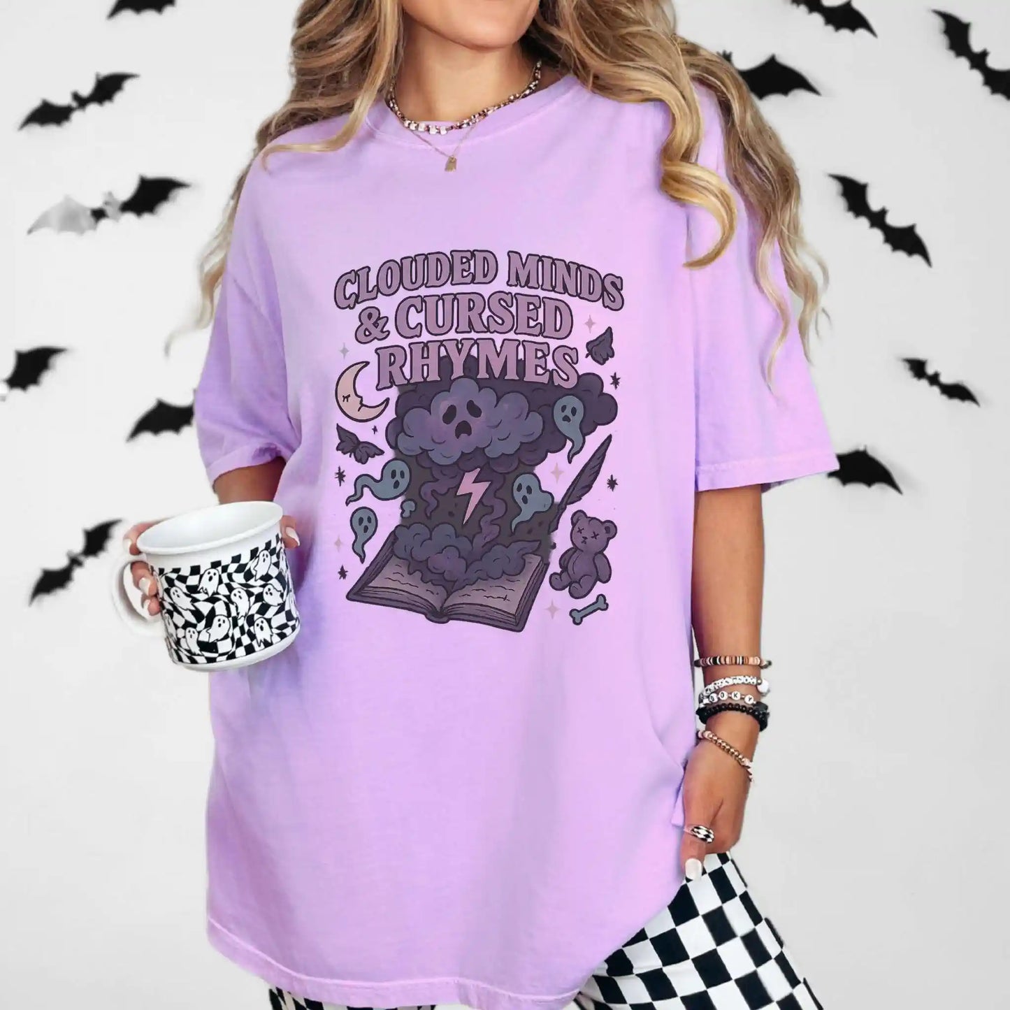 Person wearing a purple t-shirt with "clouded minds & cursed rhymes" graphic design and text, holding a mug with a Halloween-themed background.