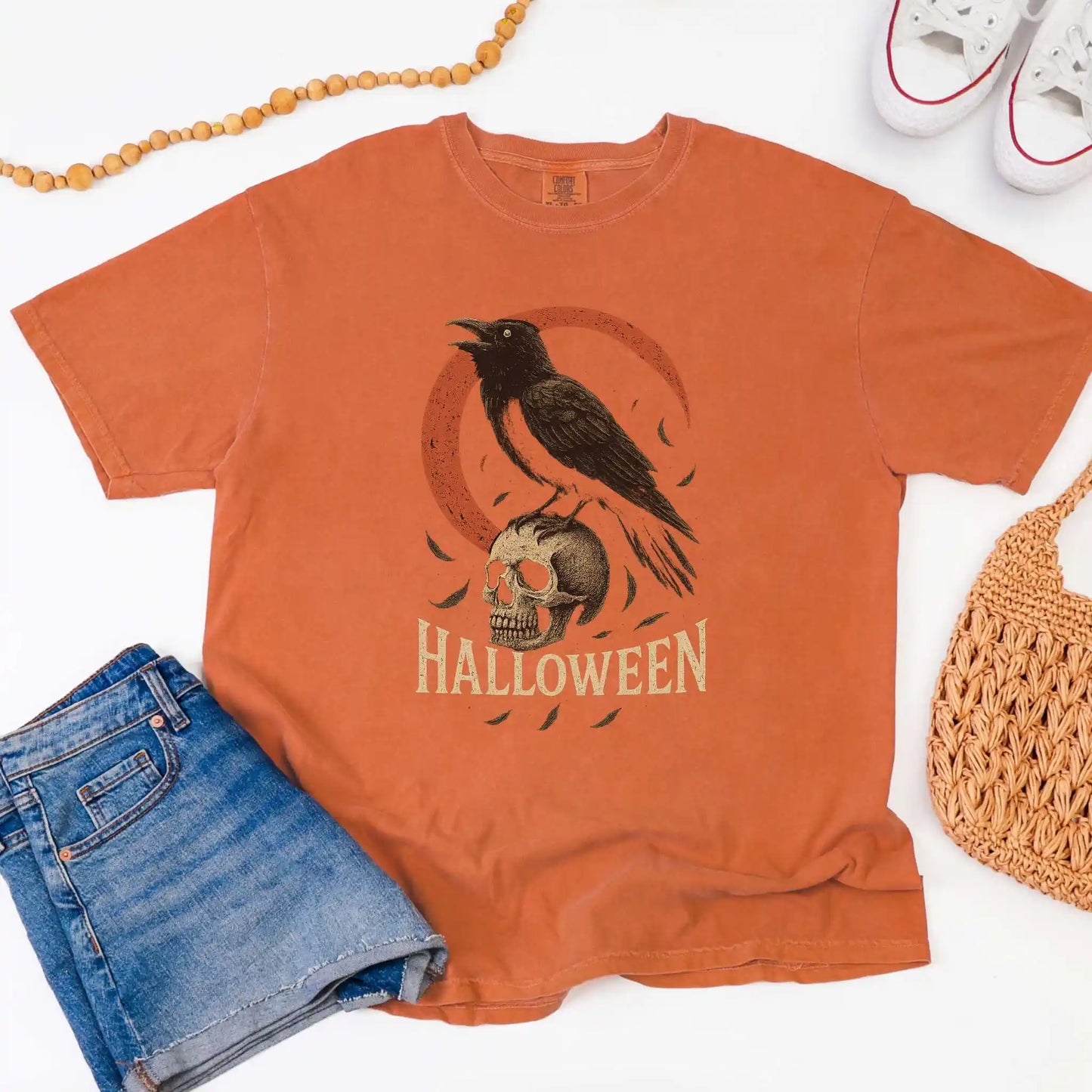 Orange t-shirt with Halloween design featuring a crow and skull, placed on a white background with jeans and a bag.