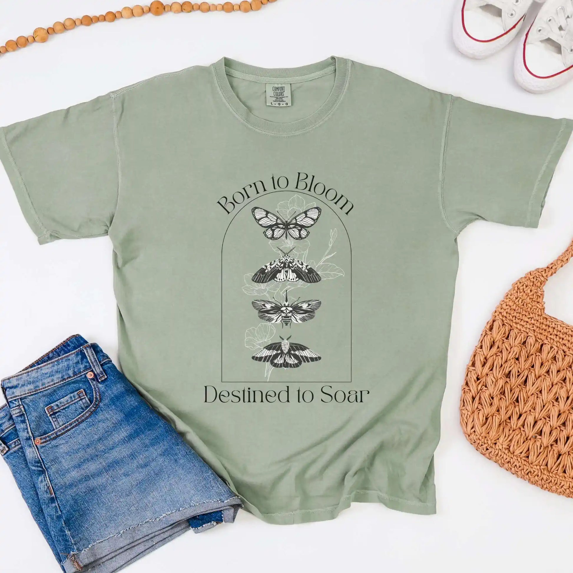 Sage green t-shirt with butterfly design and 'Born to Bloom, Destined to Soar' text, paired with jeans and a woven bag on a white background.
