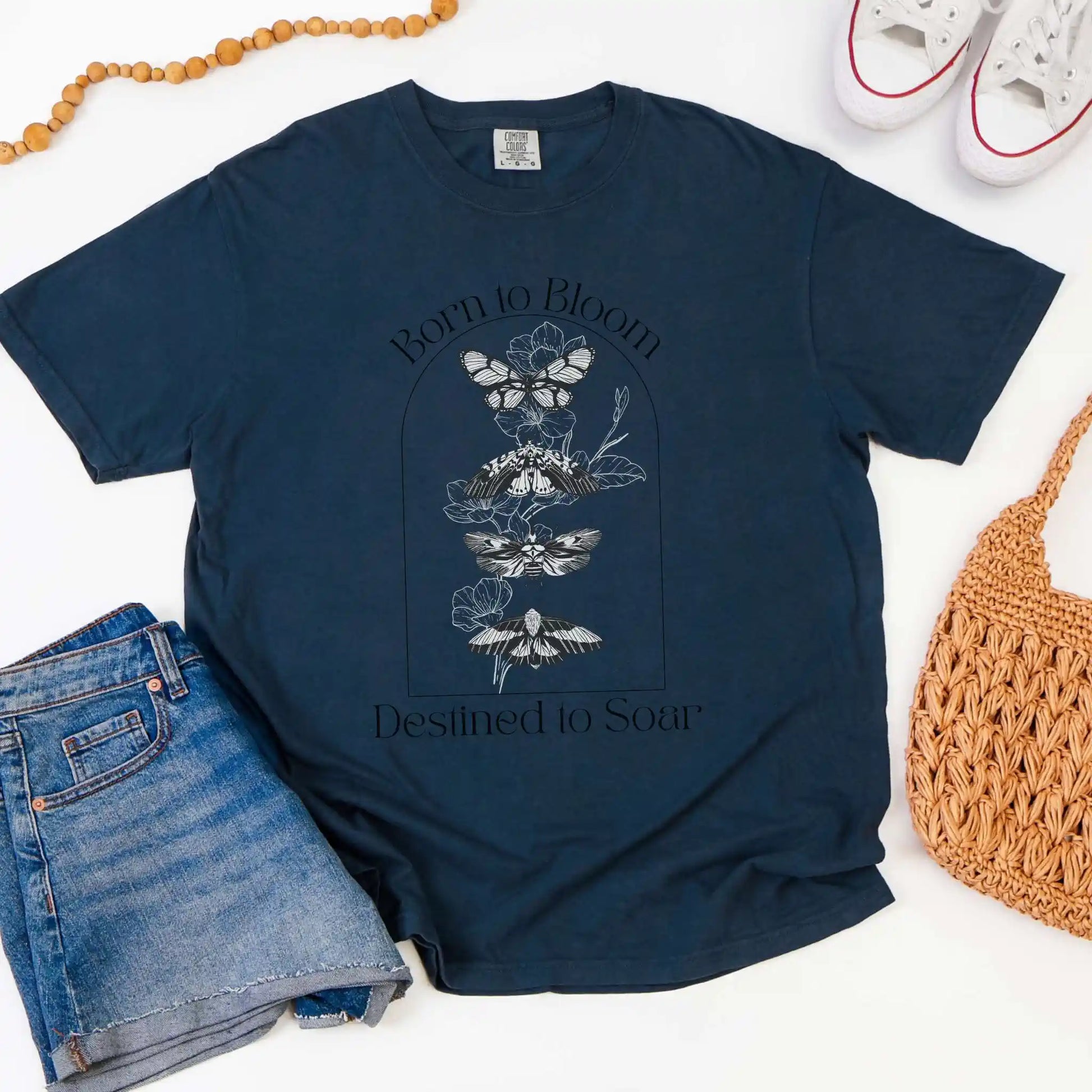 Navy blue t-shirt with butterfly design and 'Born to Bloom, Destined to Soar' text, placed on a white surface with jeans and a bag.