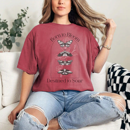 Woman wearing a red t-shirt with butterfly design and 'Born to Bloom, Destined to Soar' text, sitting on a couch.
