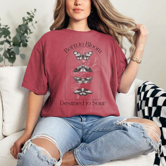 Woman wearing a red t-shirt with butterfly design and 'Born to Bloom, Destined to Soar' text, sitting on a couch.