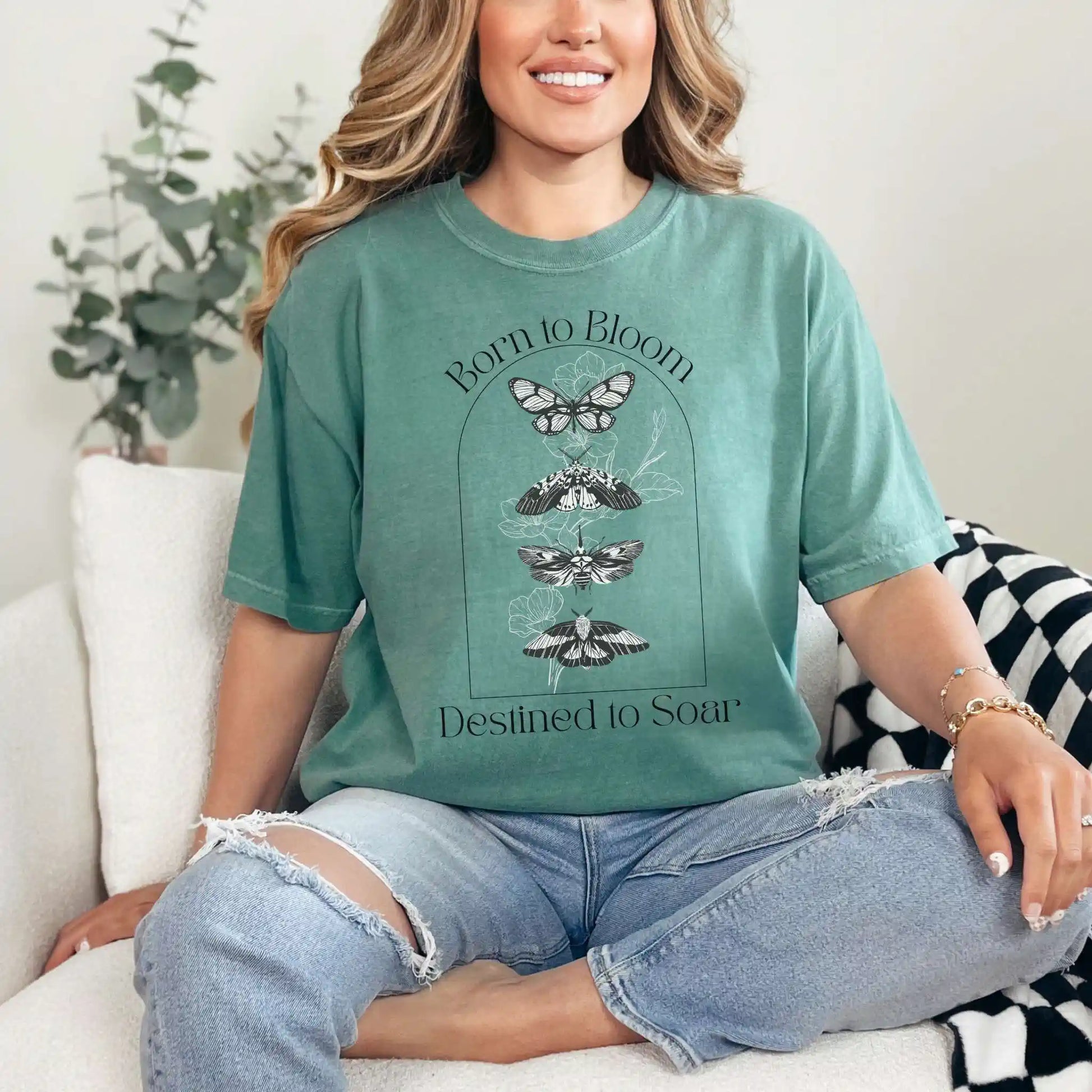 Woman wearing a light green t-shirt with butterfly design and 'Born to Bloom, Destined to Soar' text, sitting on a couch.