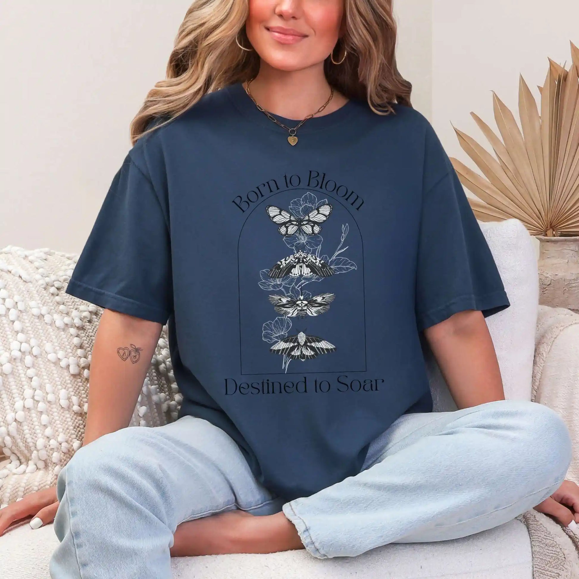 Woman wearing a navy blue t-shirt with butterfly design and 'Born to Bloom, Destined to Soar' text, sitting on a couch.