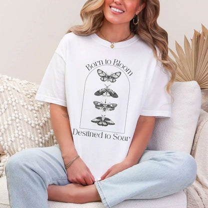 Woman wearing a white t-shirt with butterfly design and 'Born to Bloom, Destined to Soar' text, sitting on a couch.