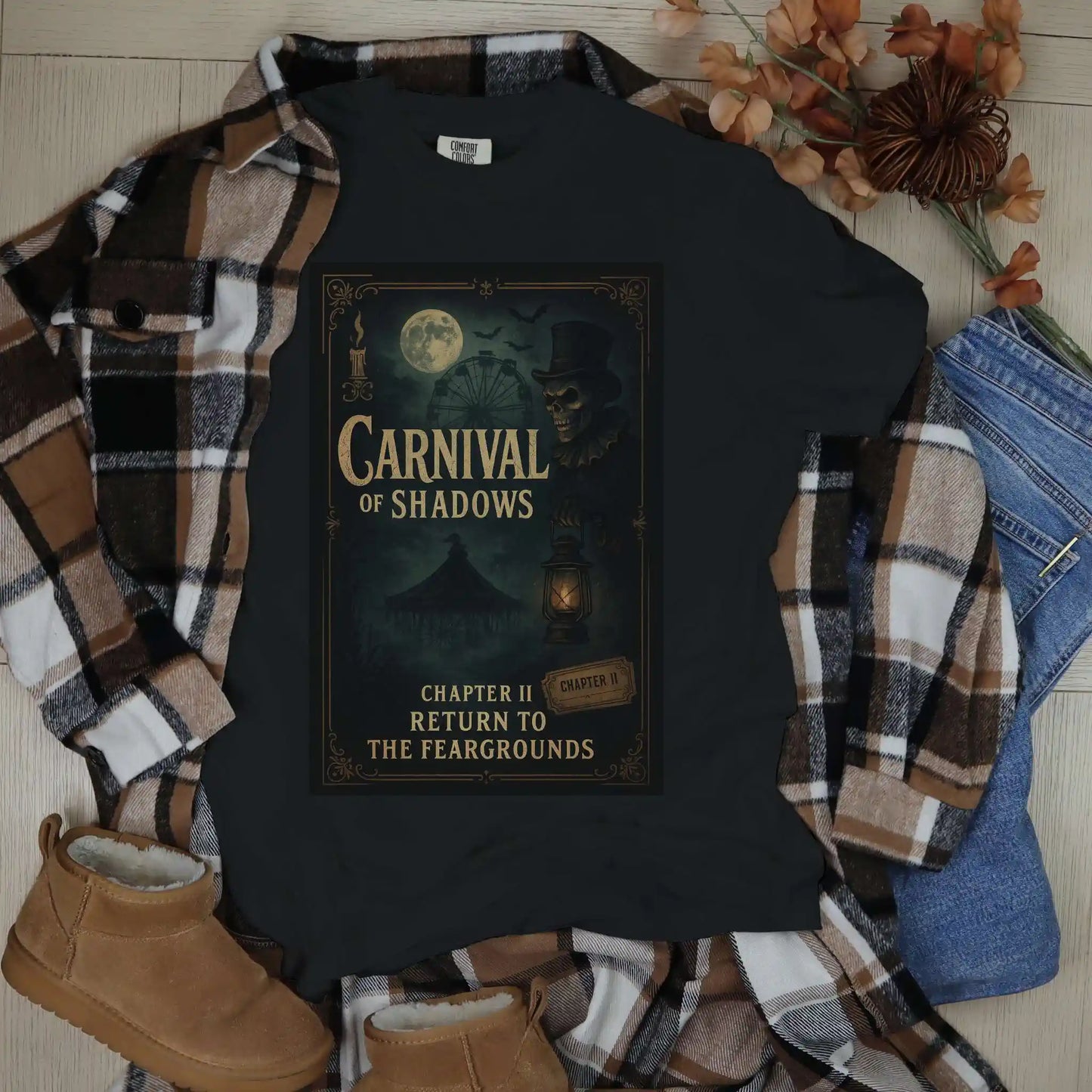 Black t-shirt with 'Carnival of Shadows' design, flannel shirt, jeans, and boots on a wooden surface.