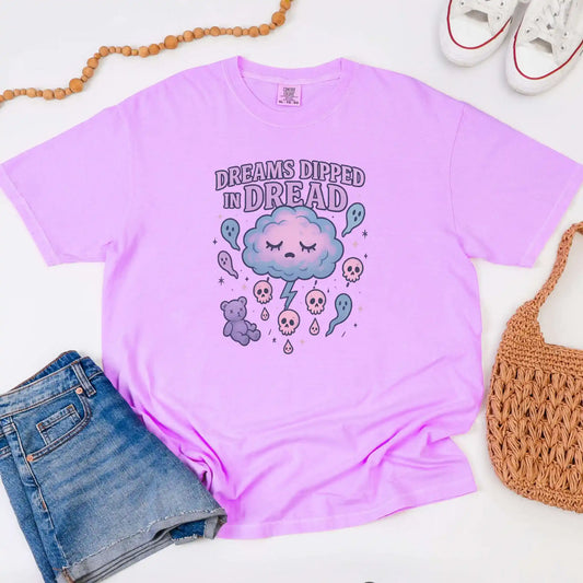 Purple t-shirt with "Dreams Dipped in Dread" graphic design with clouds and ghosts on a white background with jean shorts, and a woven bag.
