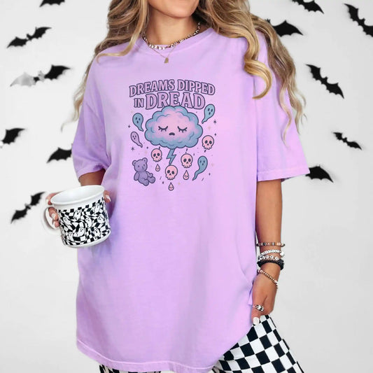 Person wearing a purple oversized t-shirt with a "Dreams Dipped in Dread" graphic design with clouds and ghosts, holding a mug with a checkered pattern, against a white background with black bats.