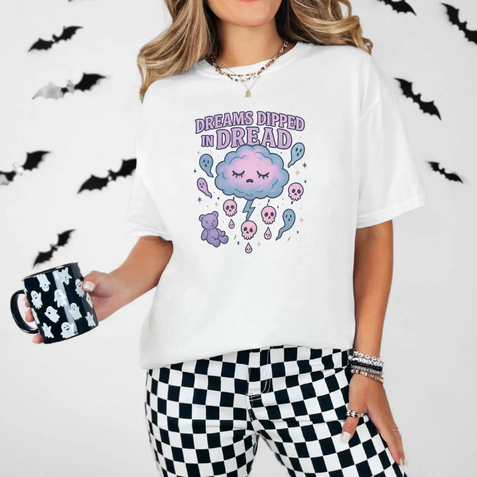 Person wearing a white t-shirt with a "Dreams Dipped in Dread" graphic design with clouds and ghosts, holding a mug, surrounded by bats.