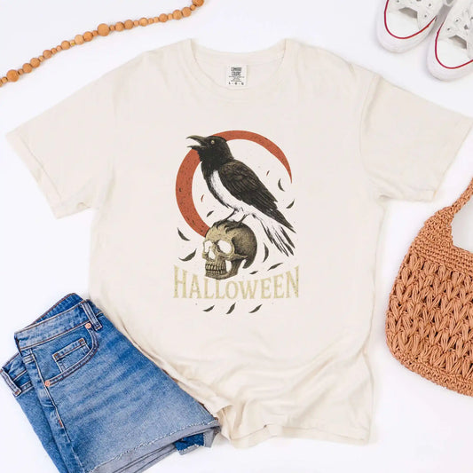Beige t-shirt with Halloween design featuring a crow and skull, paired with denim shorts and a woven bag on a white background.