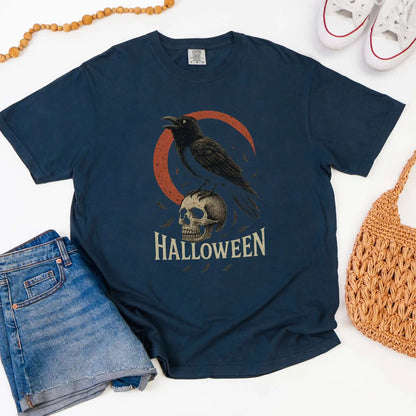 Navy blue t-shirt with Halloween design featuring a crow and skull, displayed with jeans, shoes, and a bag.