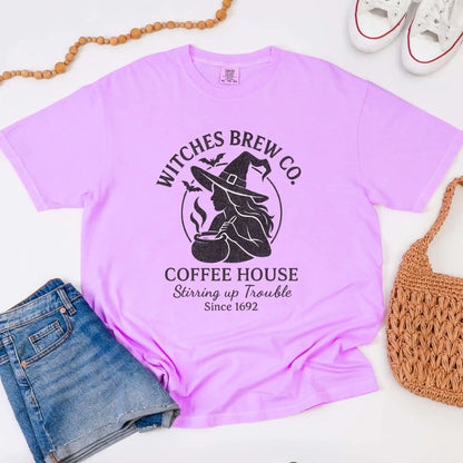 Purple t-shirt with 'Witches Brew Co. Coffee House' design on a white surface with jeans and a bag.
