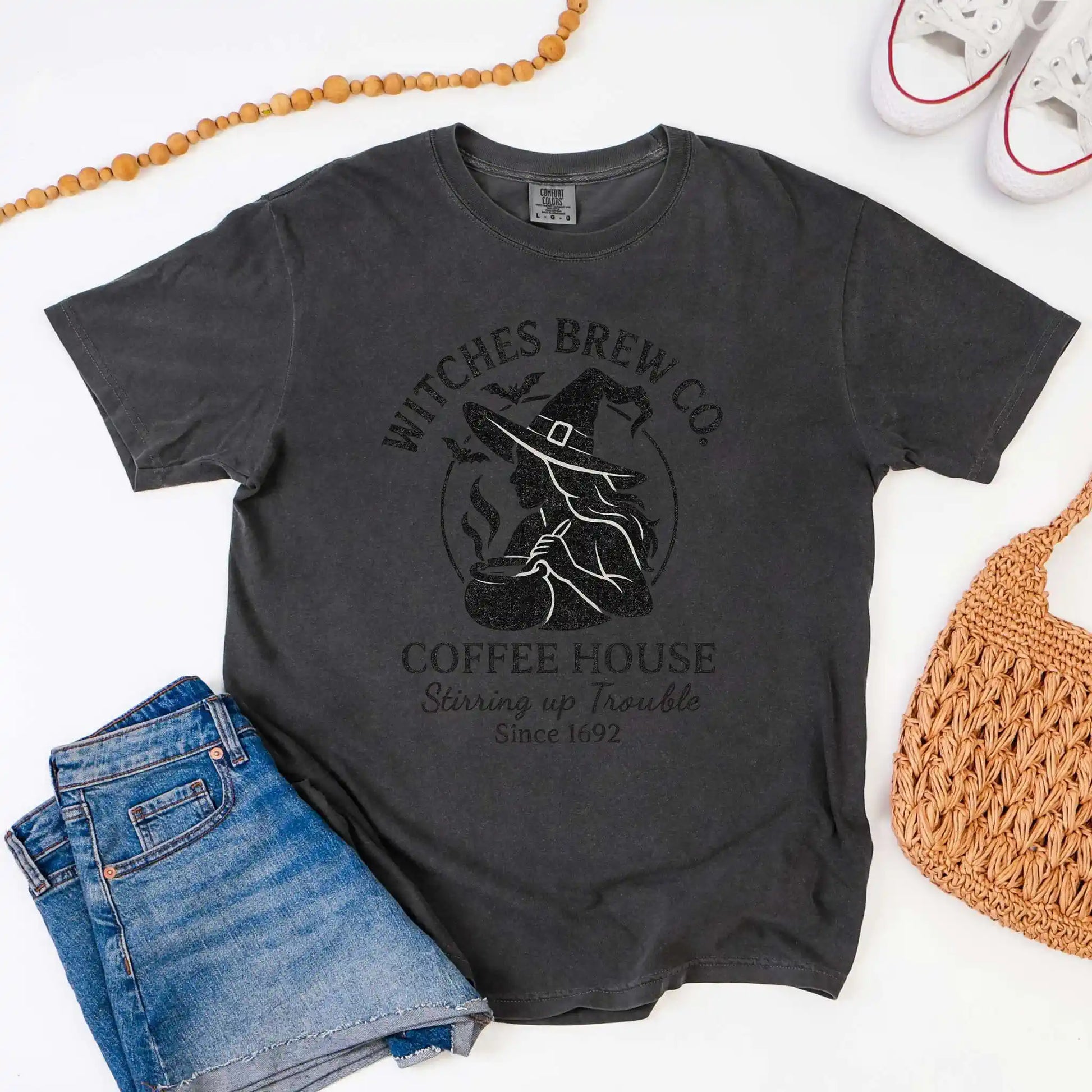 Dark gray t-shirt with 'Witches Brew Co. Coffee House' design on a white background with jeans and a bag.