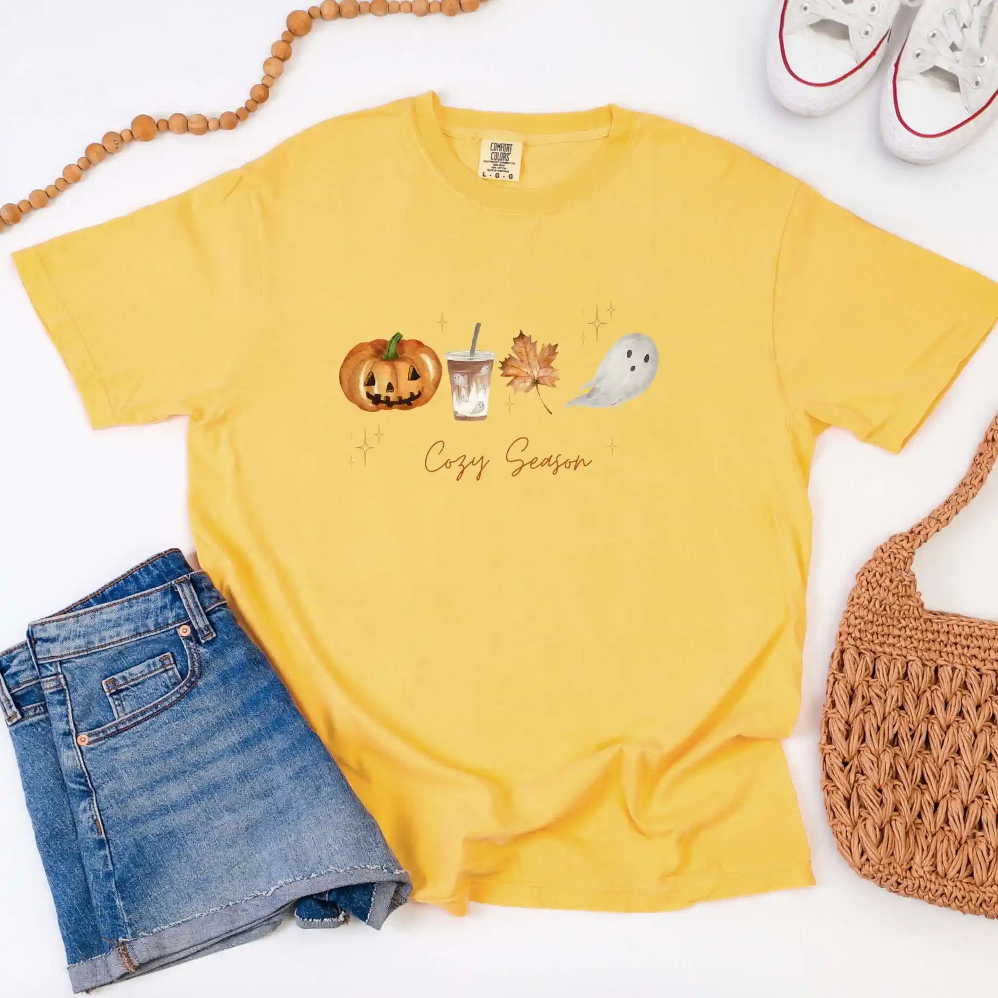 Yellow t-shirt with Halloween-themed design and 'Cozy Season' text, paired with denim shorts and a woven bag on a white background.