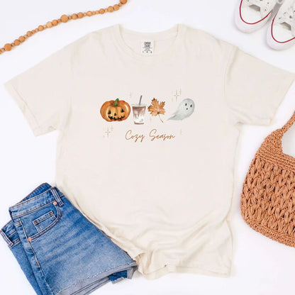 Ivory t-shirt with Halloween-themed graphics and 'Cozy Season' text, paired with denim shorts and a woven bag on a white background.