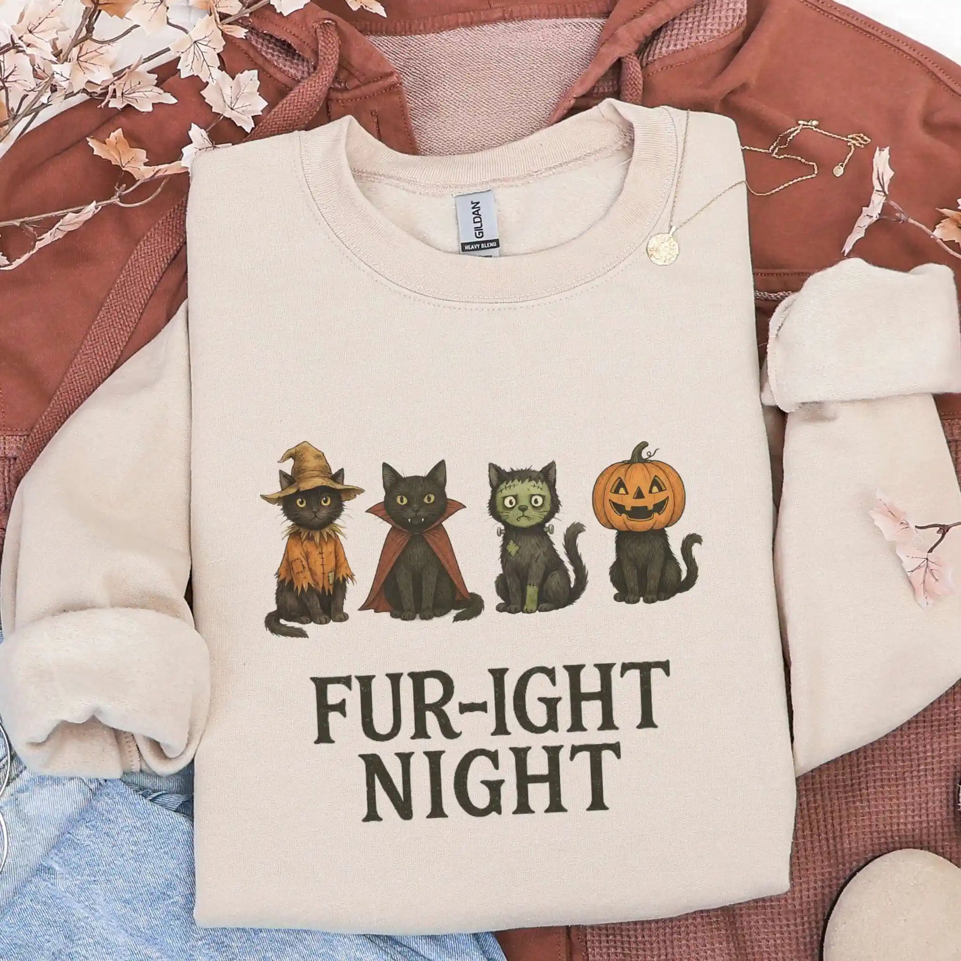 Beige sweatshirt with Halloween-themed design featuring cats in costumes and 'Fur-ight Night' text, lying on a brown jacket background.