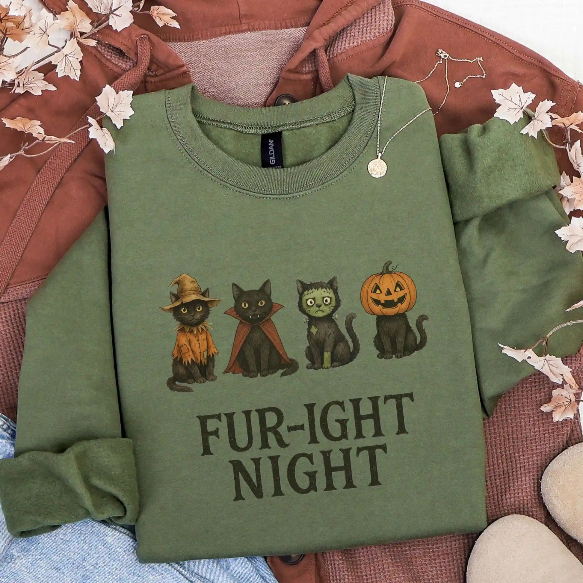Green sweatshirt with Halloween-themed design featuring cats in costumes and 'Fur-ight Night' text, lying on a brown jacket background.