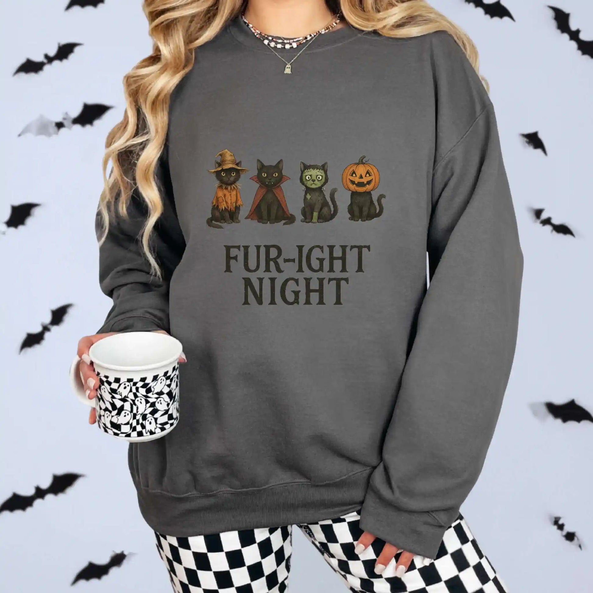A woman wearing a charcoal sweatshirt with a Halloween-themed design featuring cats in costumes and 'Fur-ight Night' text, with a white background and bat decorations
