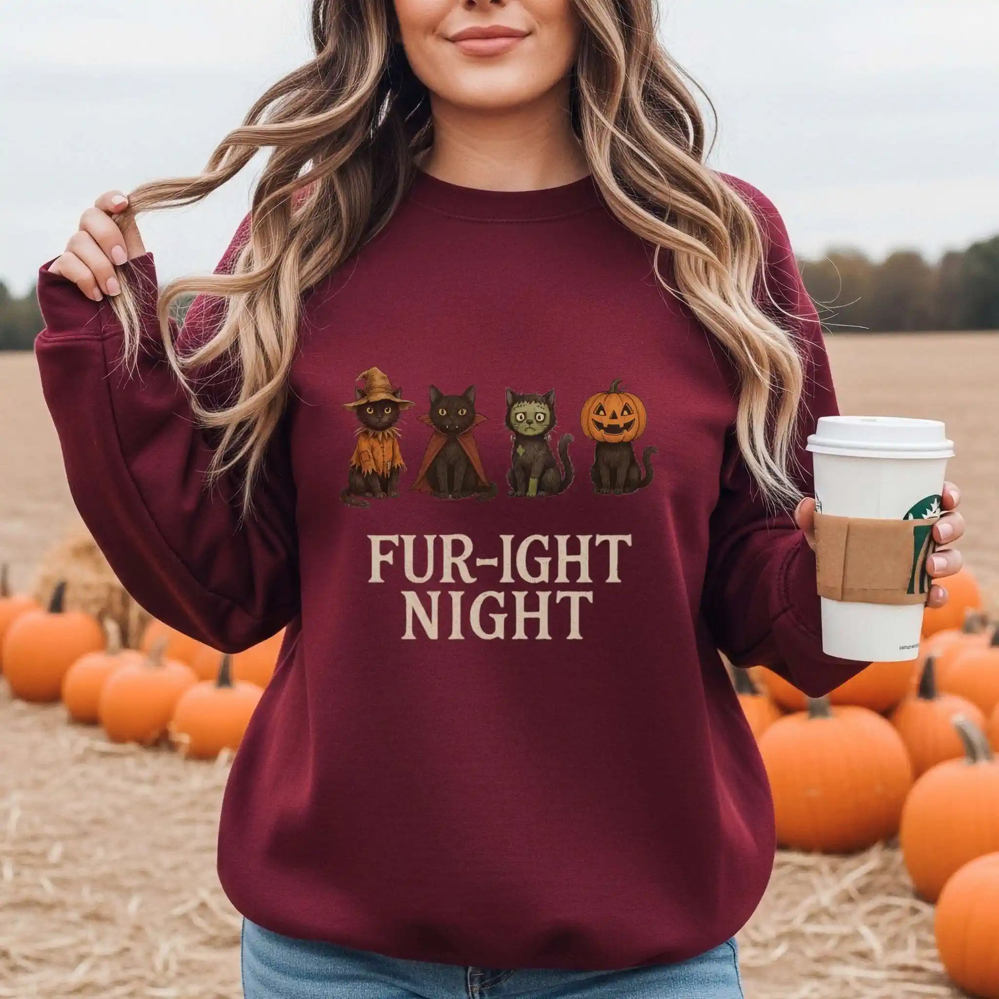 A woman wearing a maroon sweatshirt with a Halloween-themed design featuring cats in costumes and 'Fur-ight Night' text, standing in a pumpkin patch.
