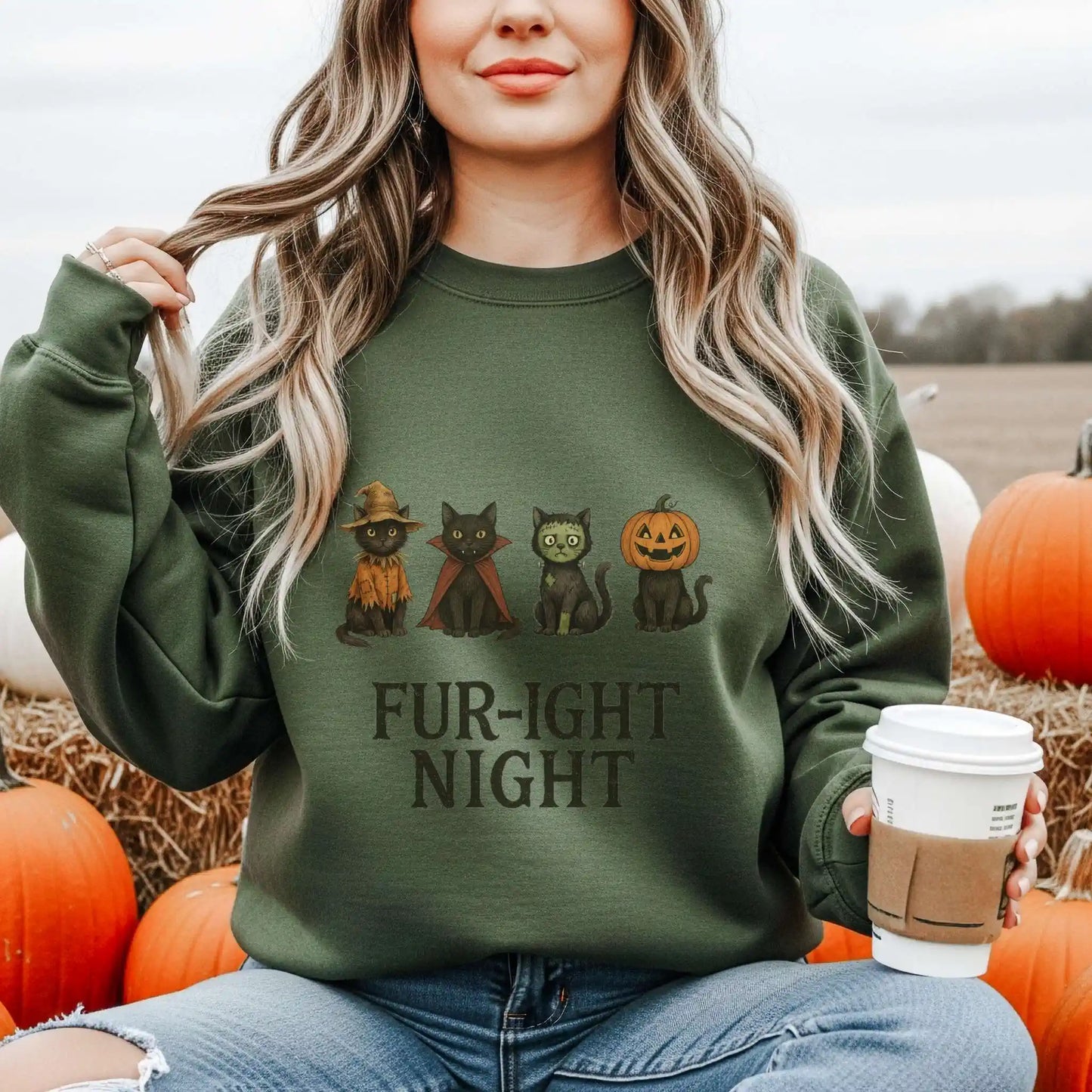 A woman wearing a military green sweatshirt with a Halloween-themed design featuring cats in costumes and 'Fur-ight Night' text, sitting in a pumpkin patch.