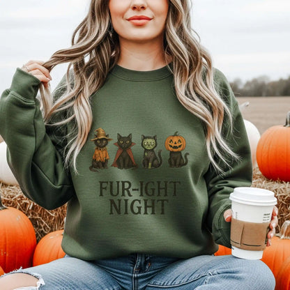 A woman wearing a military green sweatshirt with a Halloween-themed design featuring cats in costumes and 'Fur-ight Night' text, sitting in a pumpkin patch.