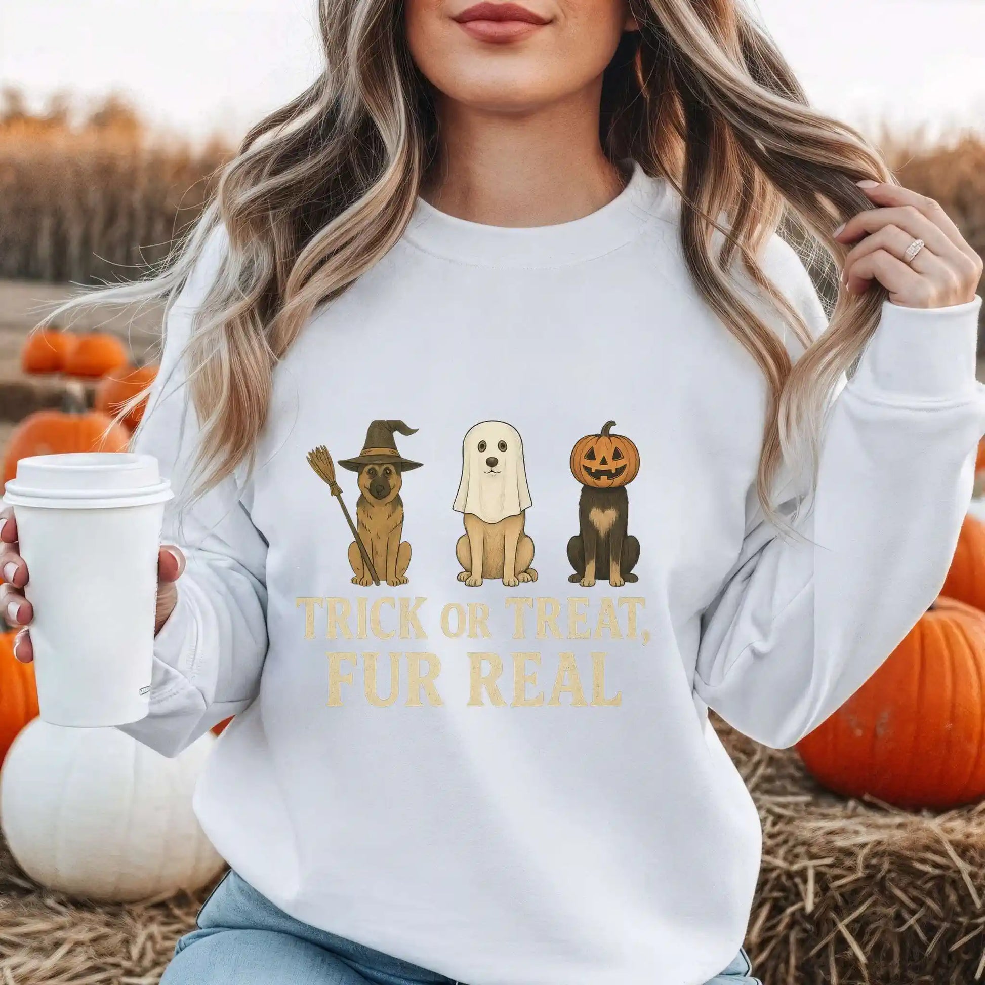 Person wearing a white sweatshirt with Dog Halloween-themed graphics and "Trick or Treat Fur Real" text, holding a coffee cup in an outdoor setting with pumpkins.