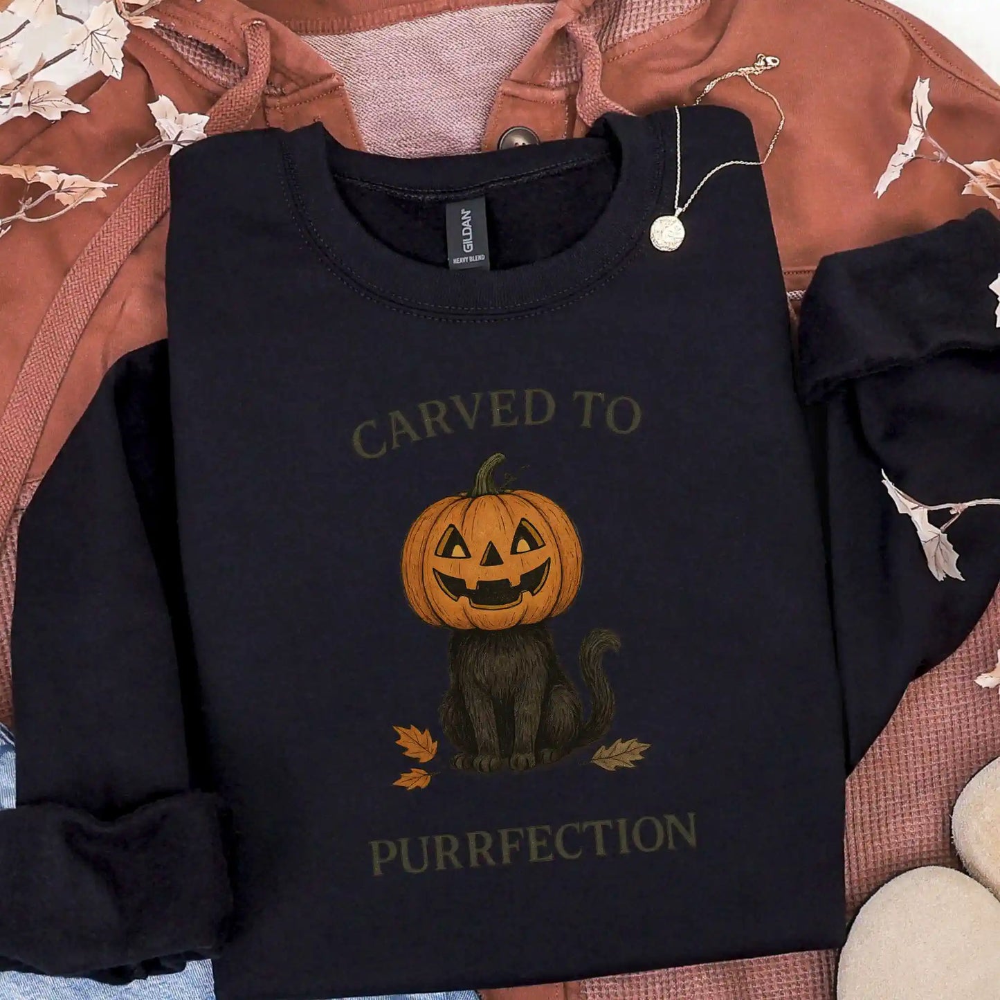 Black sweatshirt with a Halloween design of a cat with a pumpkin head and text 'Carved to Purrfection' on a brown jacket background.