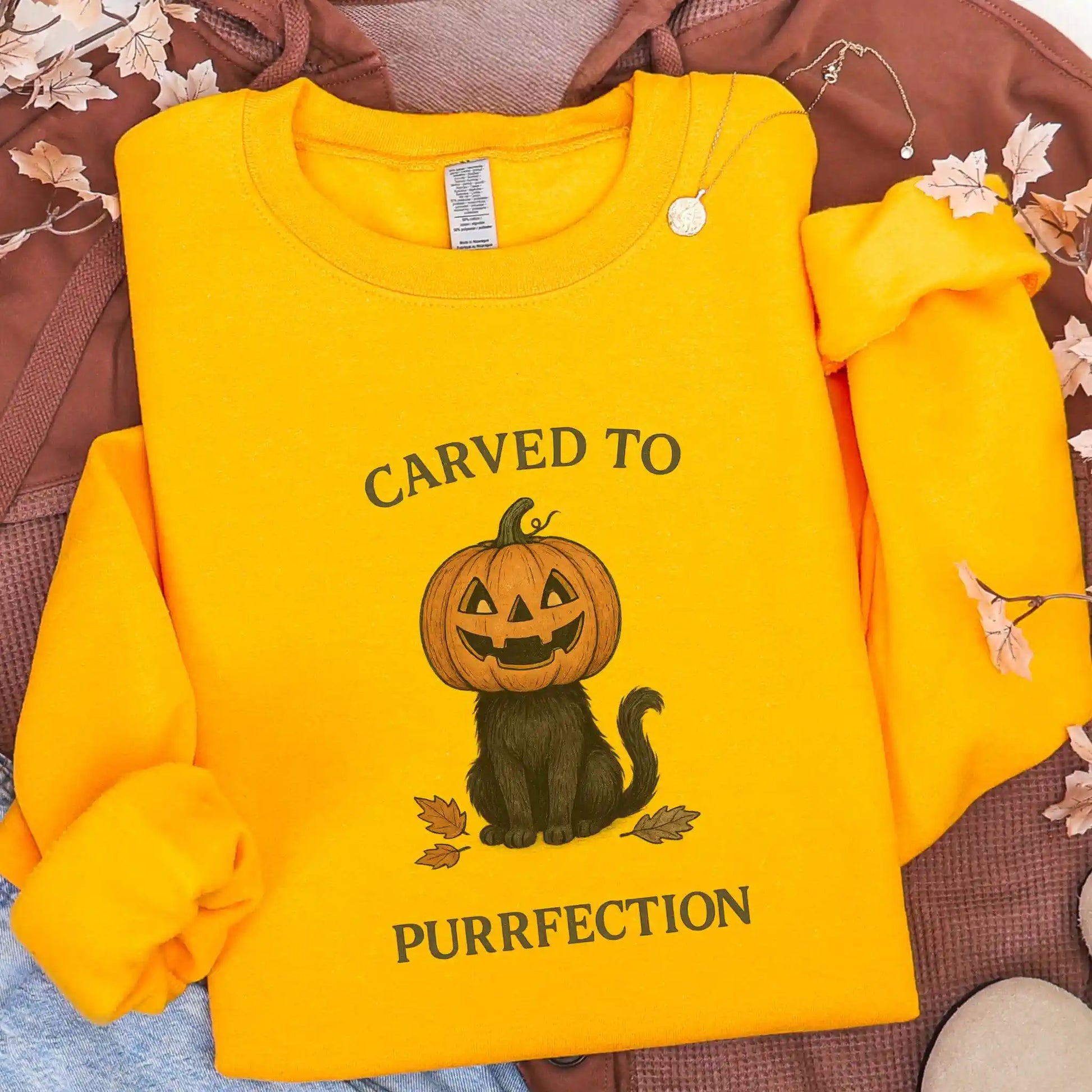Yellow sweatshirt with a Halloween design of a cat with a pumpkin head and text 'Carved to Purrfection' on a brown jacket background.