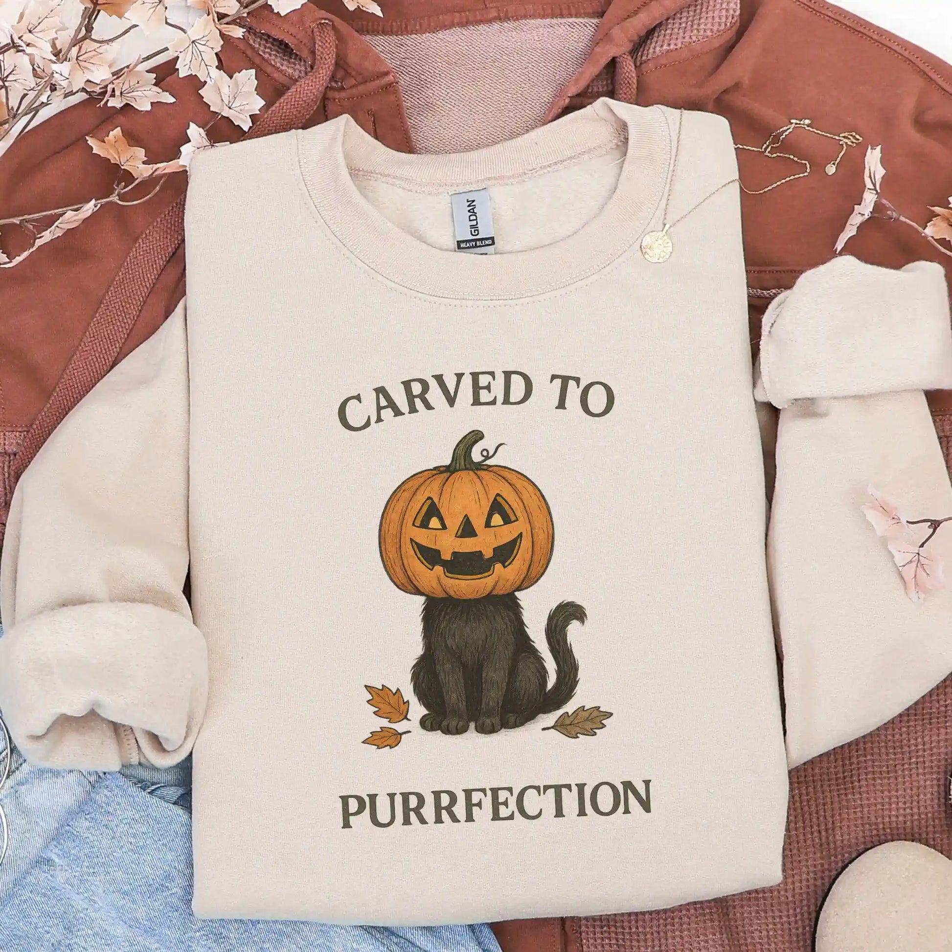 Beige sweatshirt with a Halloween design of a cat with a pumpkin head and text 'Carved to Purrfection' on a brown jacket background.