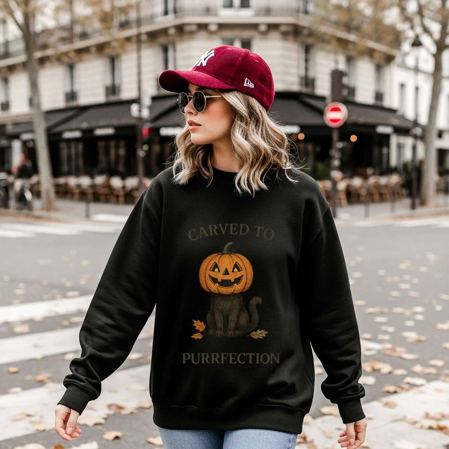A woman wearing a black sweatshirt with a Halloween design of a cat with a pumpkin head and text 'Carved to Purrfection', standing on a street.