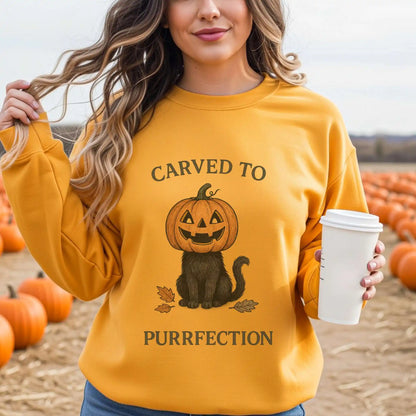 Woman wearing a yellow sweatshirt with a Halloween design of a cat with a pumpkin head and text 'Carved to Purrfection', holding a coffee cup, surrounded by pumpkins.