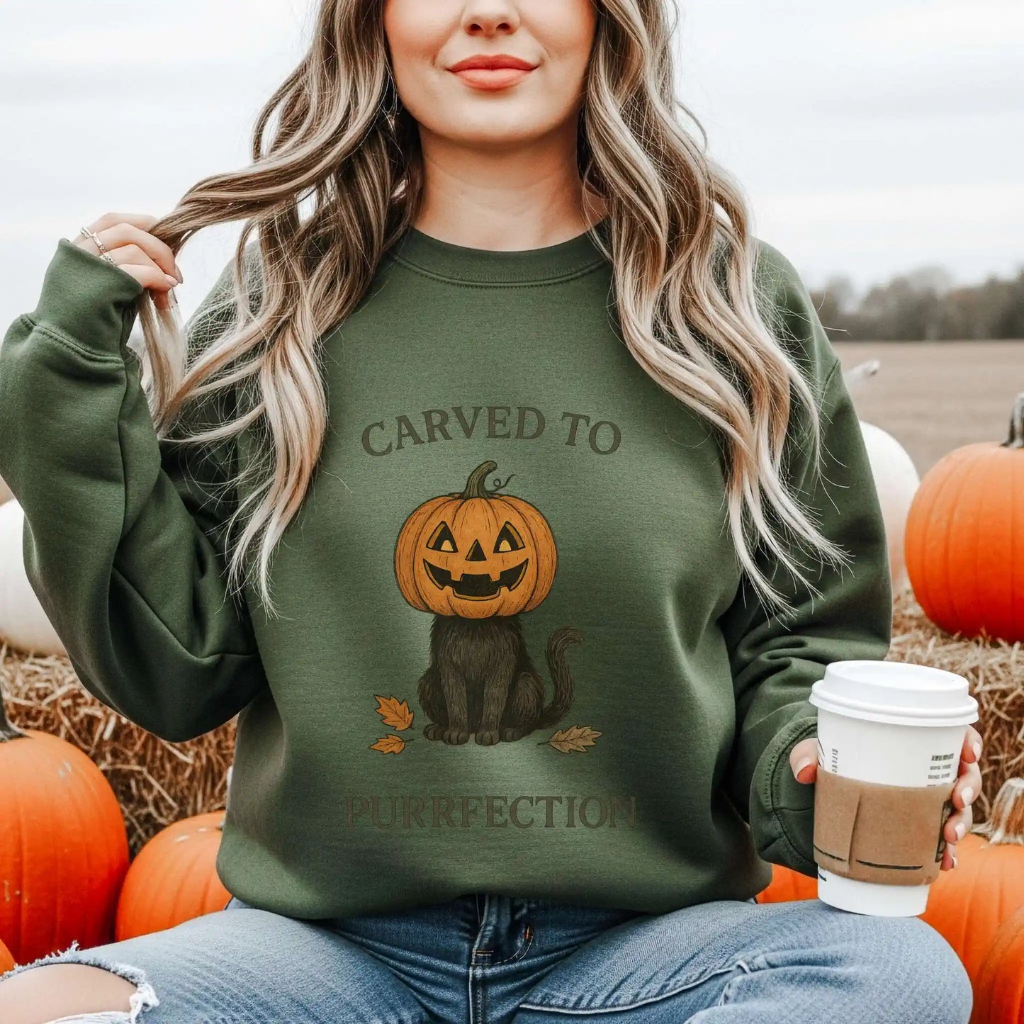 Woman wearing a green sweatshirt with a Halloween design of a cat with a pumpkin head and text 'Carved to Purrfection', holding a coffee cup, surrounded by pumpkins.