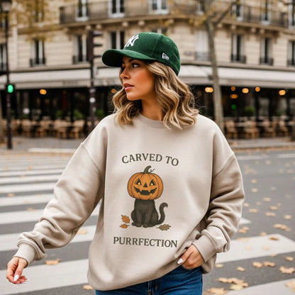A woman wearing a beige sweatshirt with a Halloween design of a cat with a pumpkin head and text 'Carved to Purrfection', standing on a street.