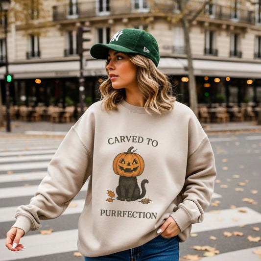 A woman wearing a beige sweatshirt with a Halloween design of a cat with a pumpkin head and text 'Carved to Purrfection', standing on a street.