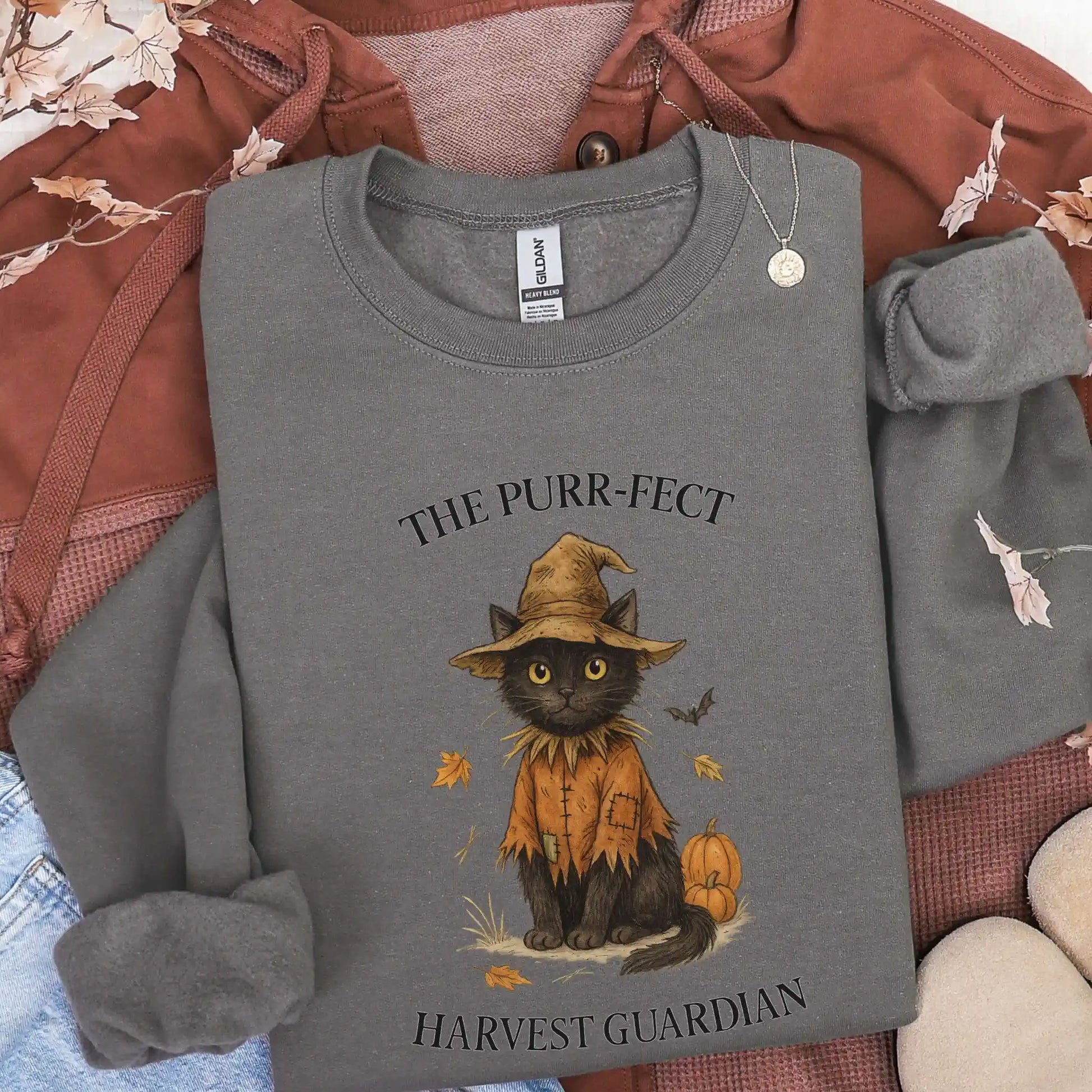Charcoal sweatshirt with a cat in a witch hat design and "The Purr-fect Harvest Guardian" text. Lying on top of a brown jacket.