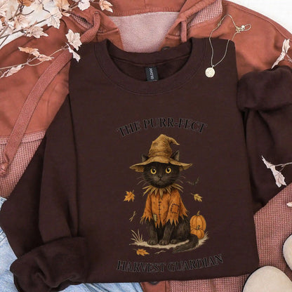 Dark Chocolate sweatshirt with a black cat in a scarecrow outfit design, with "The Purr-fect Harvest Guardian" text. Lying on a brown jacket. 
