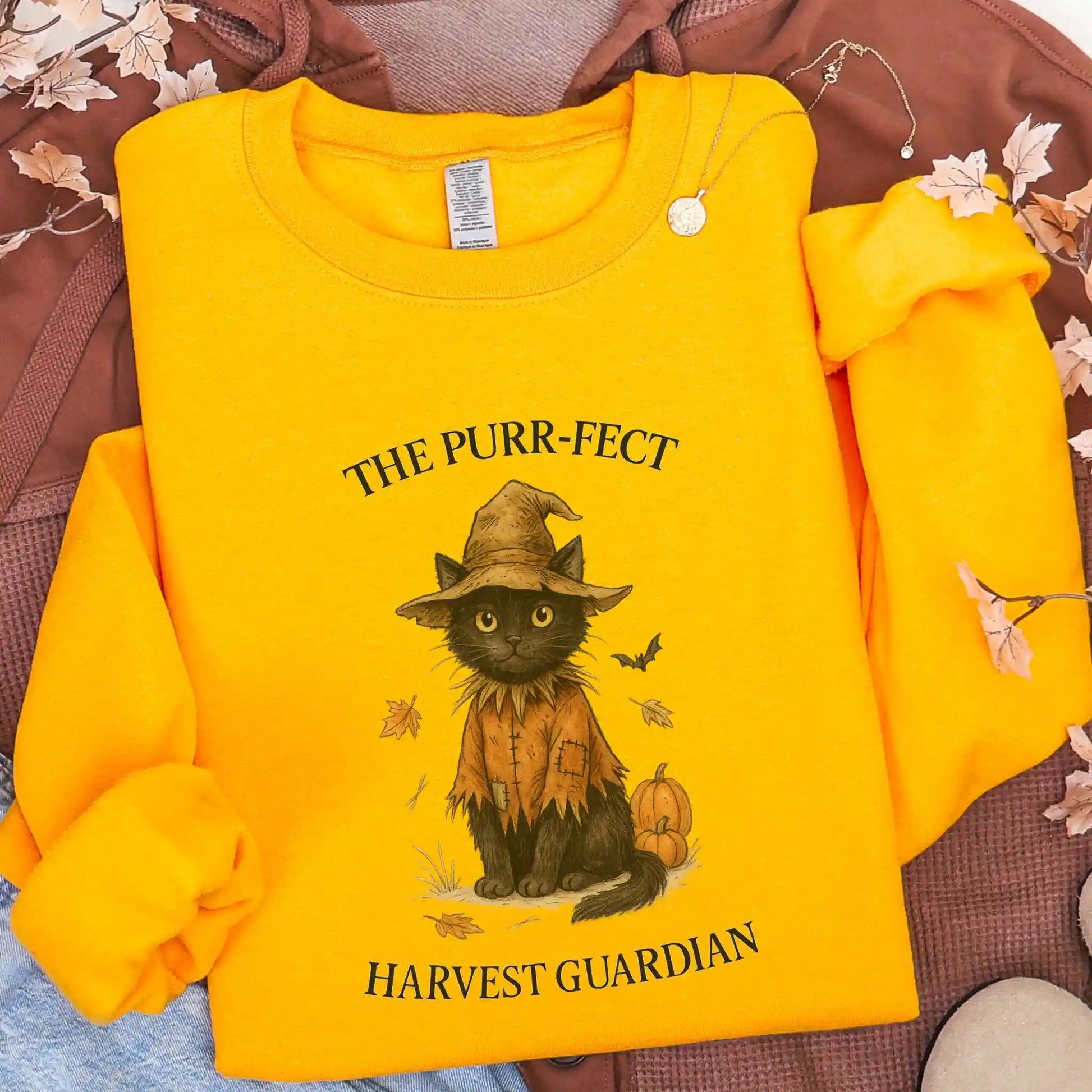 Yellow sweatshirt with a black cat in a scarecrow outfit design, with "The Purr-fect Harvest Guardian" text, lying on a brown jacket background.