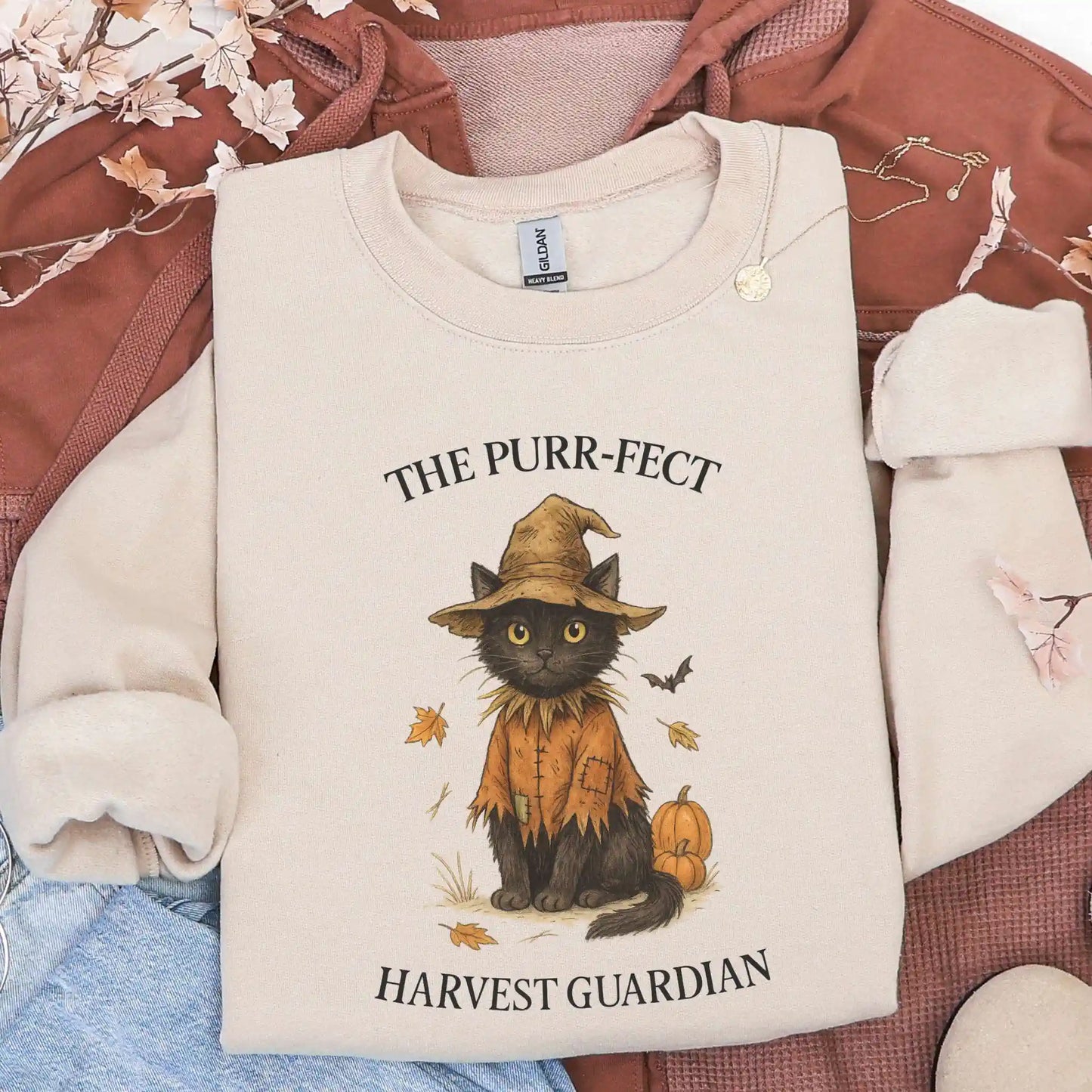 Sand colored sweatshirt with a cat wearing a tattered hat and shirt design and "The Purr-fect Harvest Guardian" text, lying on a brown jacket background.