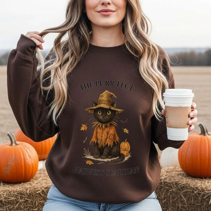 A woman wearing a brown sweatshirt with a cat in a scarecrow outfit design, with "The Purr-fect Harvest Guardian" text, holding a coffee cup in a pumpkin patch.