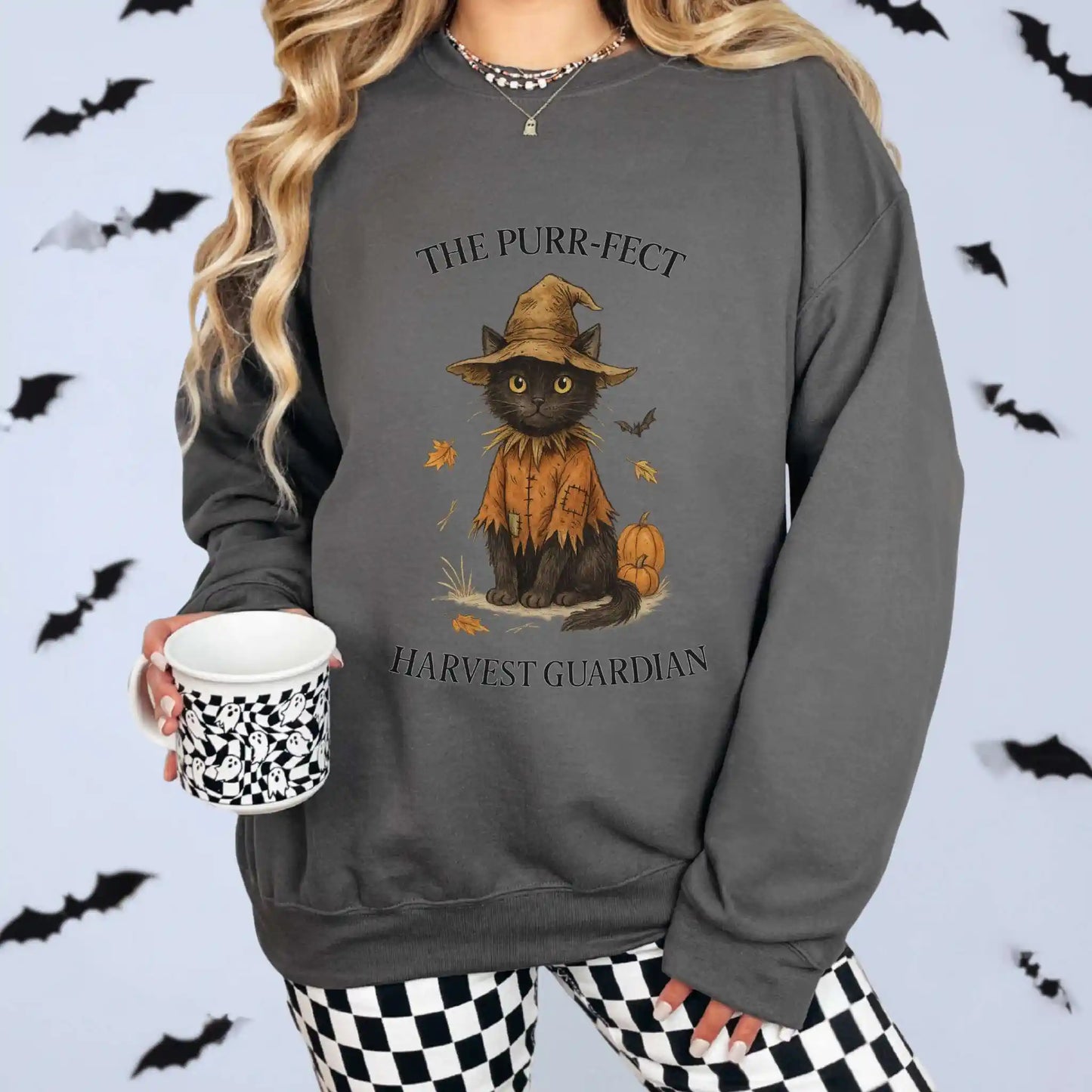 A woman wearing a charcoal sweatshirt with a cat in an orange outfit and hat design, surrounded by Halloween-themed decorations. "The Purr-fect Harvest Guardian" text around the cat. Bats in the background for Halloween feel.