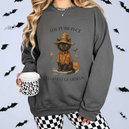 A woman wearing a charcoal sweatshirt with a cat in an orange outfit and hat design, surrounded by Halloween-themed decorations. "The Purr-fect Harvest Guardian" text around the cat. Bats in the background for Halloween feel.