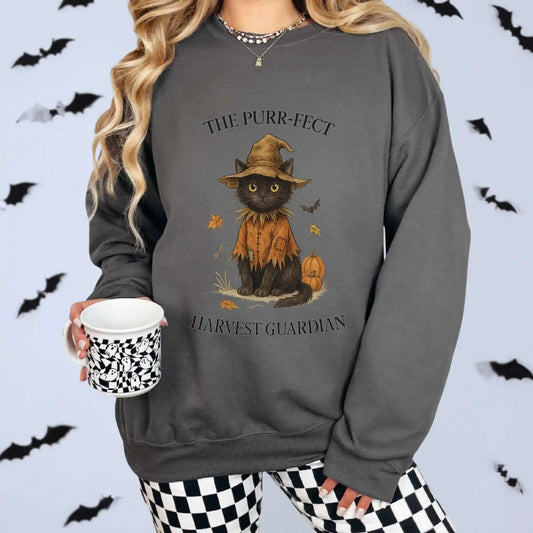A woman wearing a charcoal sweatshirt with a cat in an orange outfit and hat design, surrounded by Halloween-themed decorations. "The Purr-fect Harvest Guardian" text around the cat. Bats in the background for Halloween feel.