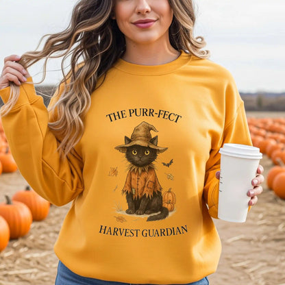 Woman wearing a yellow sweatshirt with a cat in a scarecrow outfit design, with "The Purr-fect Harvest Guardian" text, holding a coffee cup in a pumpkin patch.