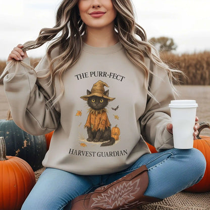 A woman wearing a Sand colored sweatshirt with a cat in a scarecrow costume graphic, "The Purr-fect Harvest Guardian" text, sitting among pumpkins in a field.