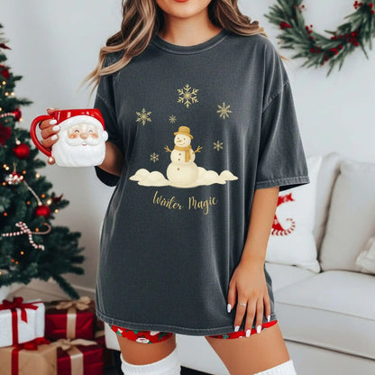 A woman wearing a pepper (dark grey) colored t-shirt with a snowman design and 'Winter Magic' text, holding a Santa mug in a festive room.