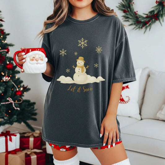 A woman wearing a dark pepper (grey) colored t-shirt with a snowman design with text 'Let it Snow', holding a Santa mug, in a festive living room.