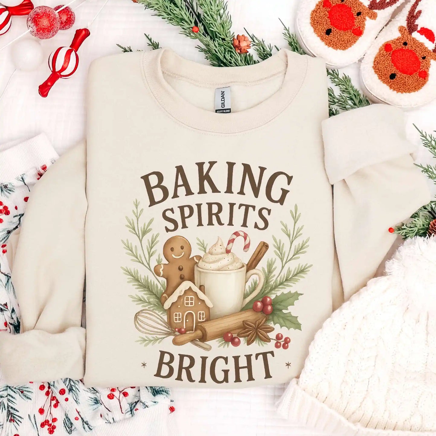 Creamy sand-colored Beacon & Bramble Christmas sweatshirt featuring the phrase ‘Baking Spirits Bright’ above a festive design with a gingerbread man, mug of cocoa topped with whipped cream and a candy cane, cinnamon sticks, greenery, and holly berries. Surrounded by cozy holiday decor including a knit hat, reindeer slippers, and greenery accents.