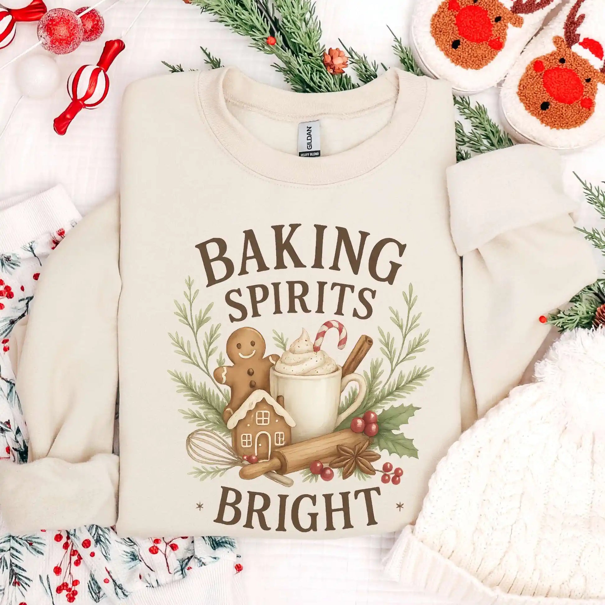 Creamy sand-colored Beacon & Bramble Christmas sweatshirt featuring the phrase ‘Baking Spirits Bright’ above a festive design with a gingerbread man, mug of cocoa topped with whipped cream and a candy cane, cinnamon sticks, greenery, and holly berries. Surrounded by cozy holiday decor including a knit hat, reindeer slippers, and greenery accents.